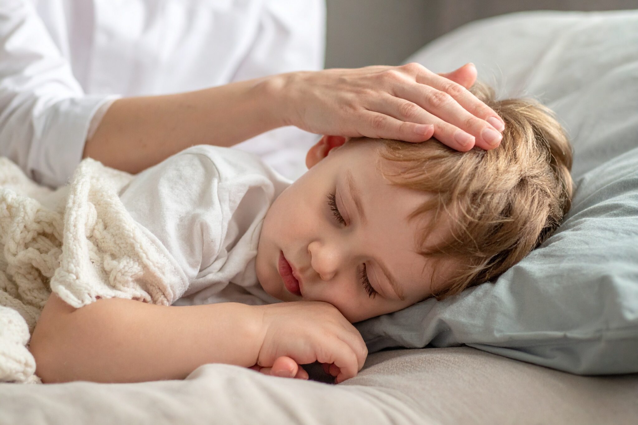 Ein kleiner blonder Junge liegt schlafend und mit einer beigen Decke zugedeckt im Bett. Der Kopf ruht auf einem grauen Polster und jemand legt sanft seine Hand auf dessen Kopf.