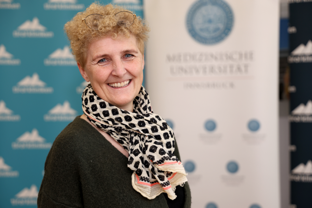 Eine lächelnde Frau mittleren Alters mit kurzen, lockigen, hellblonden Haaren blickt in die Kamera. Sie trägt eine Brille auf dem Kopf, einen dunkelgrünen Pullover und einen schwarz-weiß gemusterten Schal. Der Hintergrund ist unscharf und zeigt Roll-up-Banner mit Logos und Schriftzügen, unter anderem einer medizinischen Universität. Die Atmosphäre wirkt freundlich und professionell.