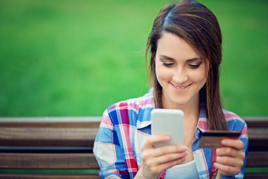 Mädchen sitzt auf einer Parkband und machen eine Onlineüberweisung am Handy, in der anderen Hand hält sie die Bankomatkarte